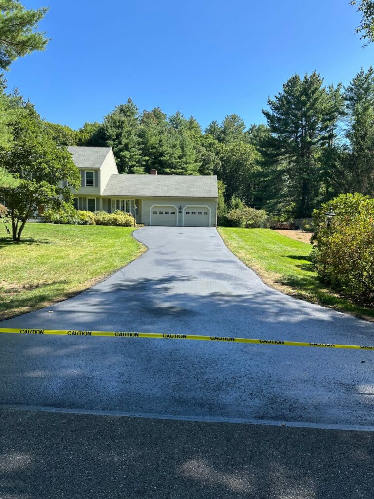 Freshly sealcoated driveway shaded by tall pines, leading to a two-car garage in Bolton, MA.